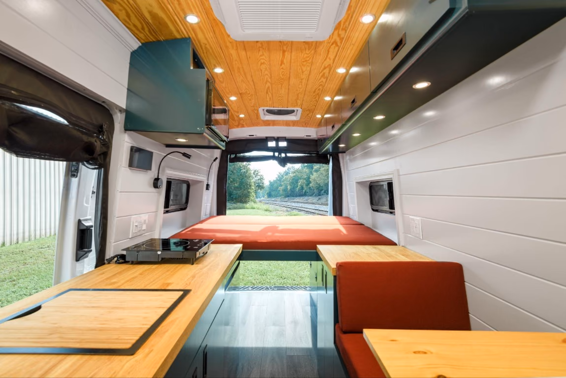 Fixed bed at the rear of a camper van with a view through the open back doors, tinted bunk windows, and storage below.