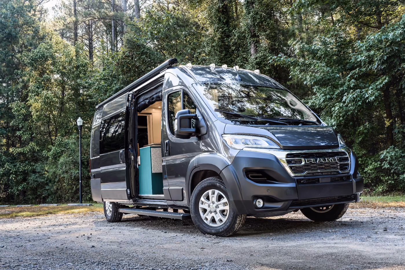 Custom RAM ProMaster camper van conversion by Crescent Moon Vans with a retractable awning, side running boards, and an open sliding door parked among trees.